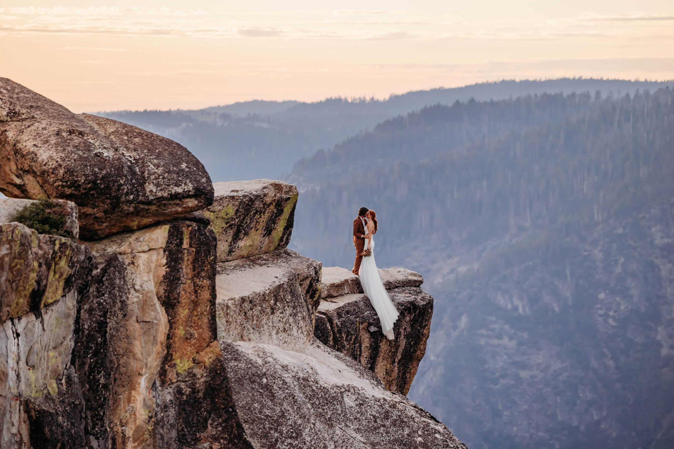 Bride and groom standing on the edge of a cliff at Taft Point during their Yosemite elopement, kissing at sunset with mountain views in the background.