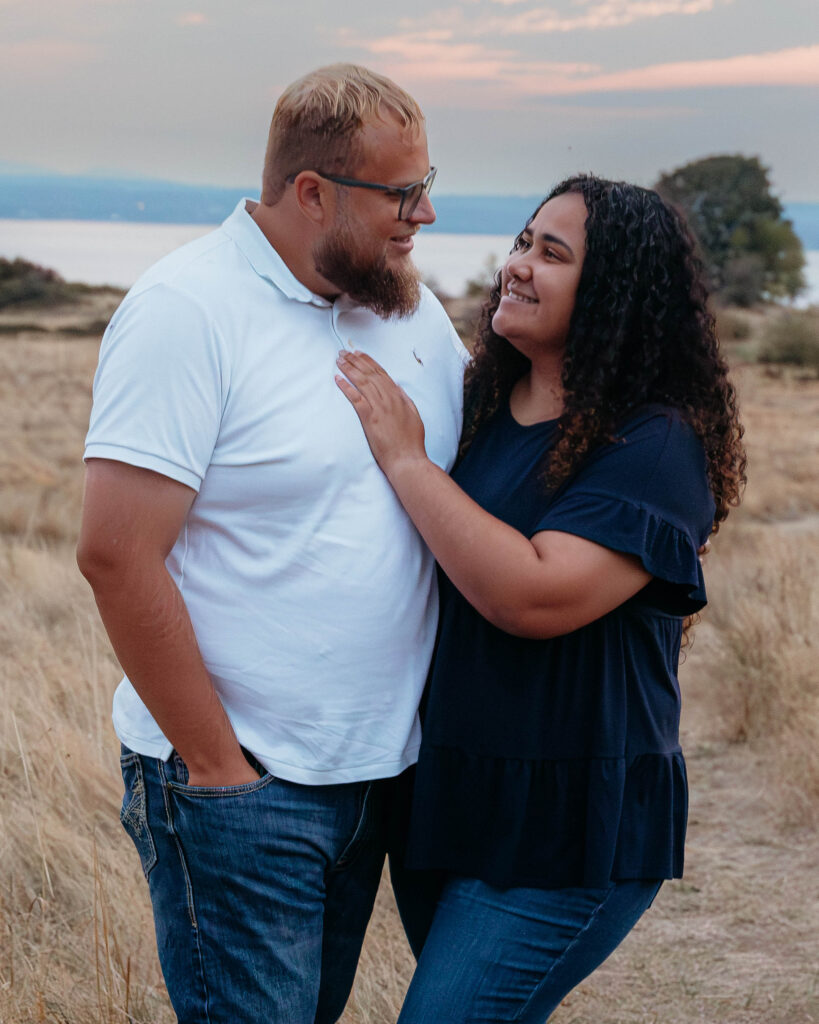 Interracial couple smiling and standing together in a golden field at Discovery Park in Seattle during sunset.