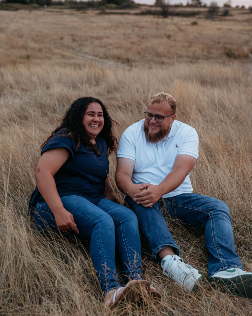 Interracial couple sitting close in the grass with Puget Sound behind them at Discovery Park during sunset.