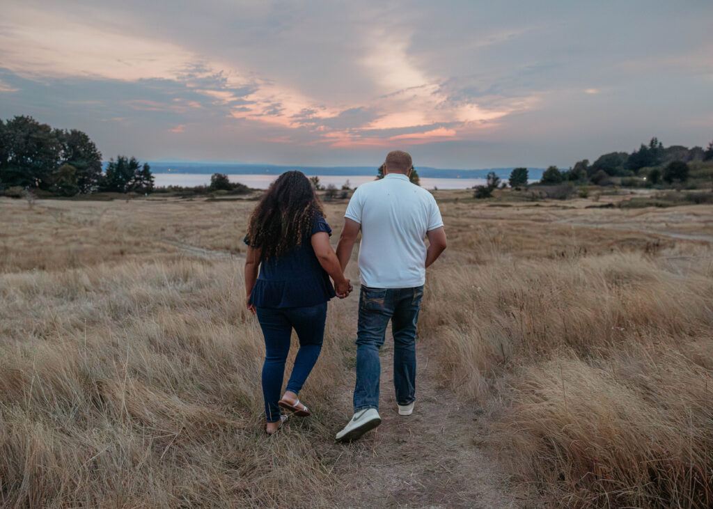 Interracial couple walking through a golden meadow at Discovery Park in Seattle with soft sunset colors in the sky.