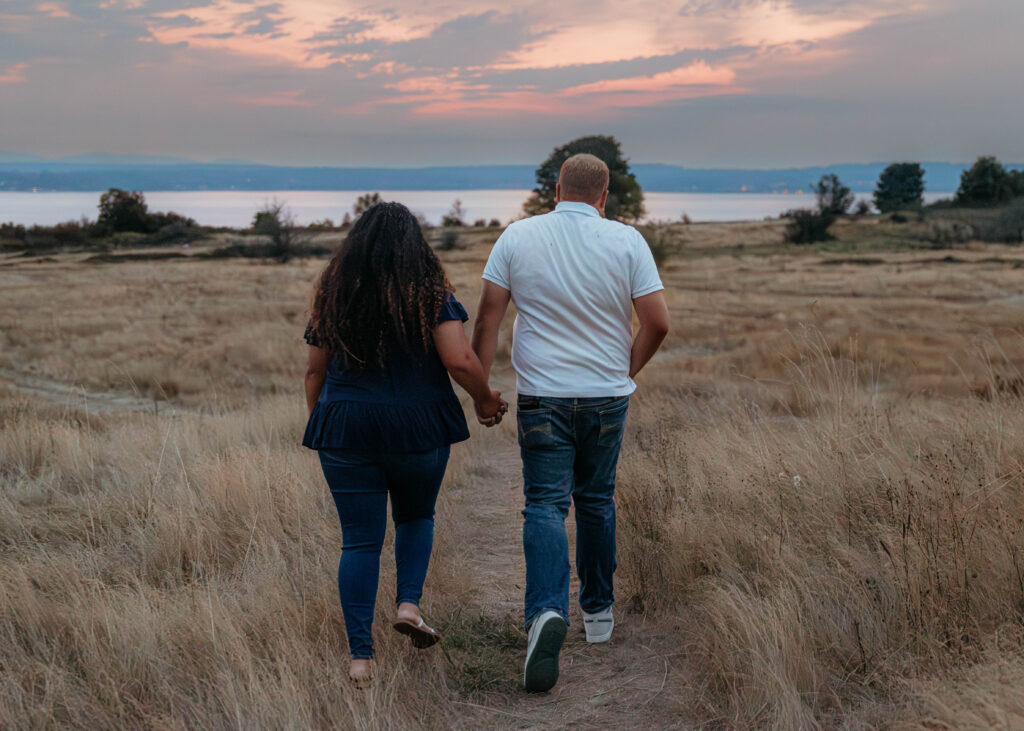Interracial couple holding hands while walking through tall golden grass toward the sunset at Discovery Park in Seattle.
