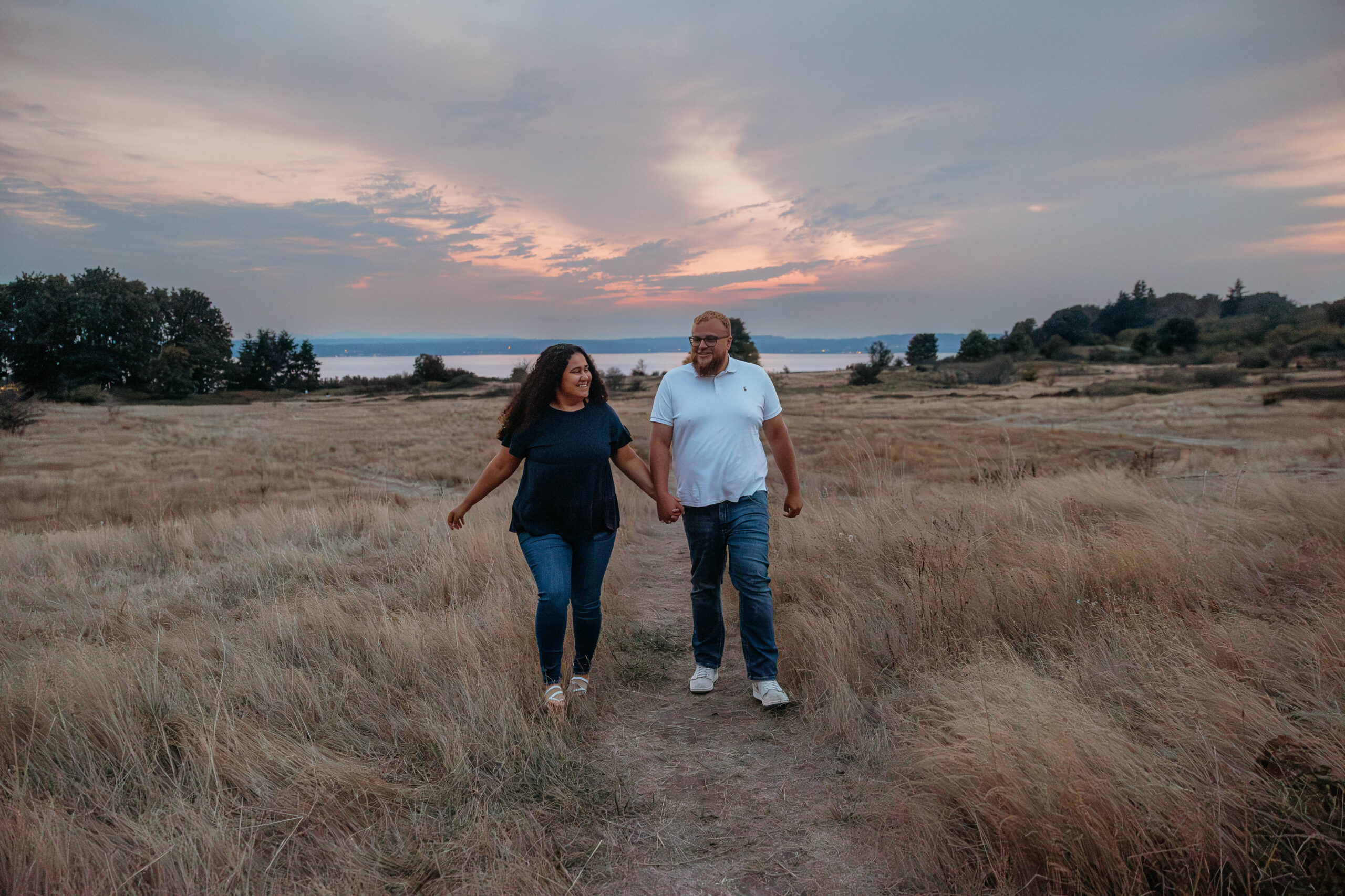 Interracial couple sitting close in the grass with Puget Sound behind them at Discovery Park during sunset.